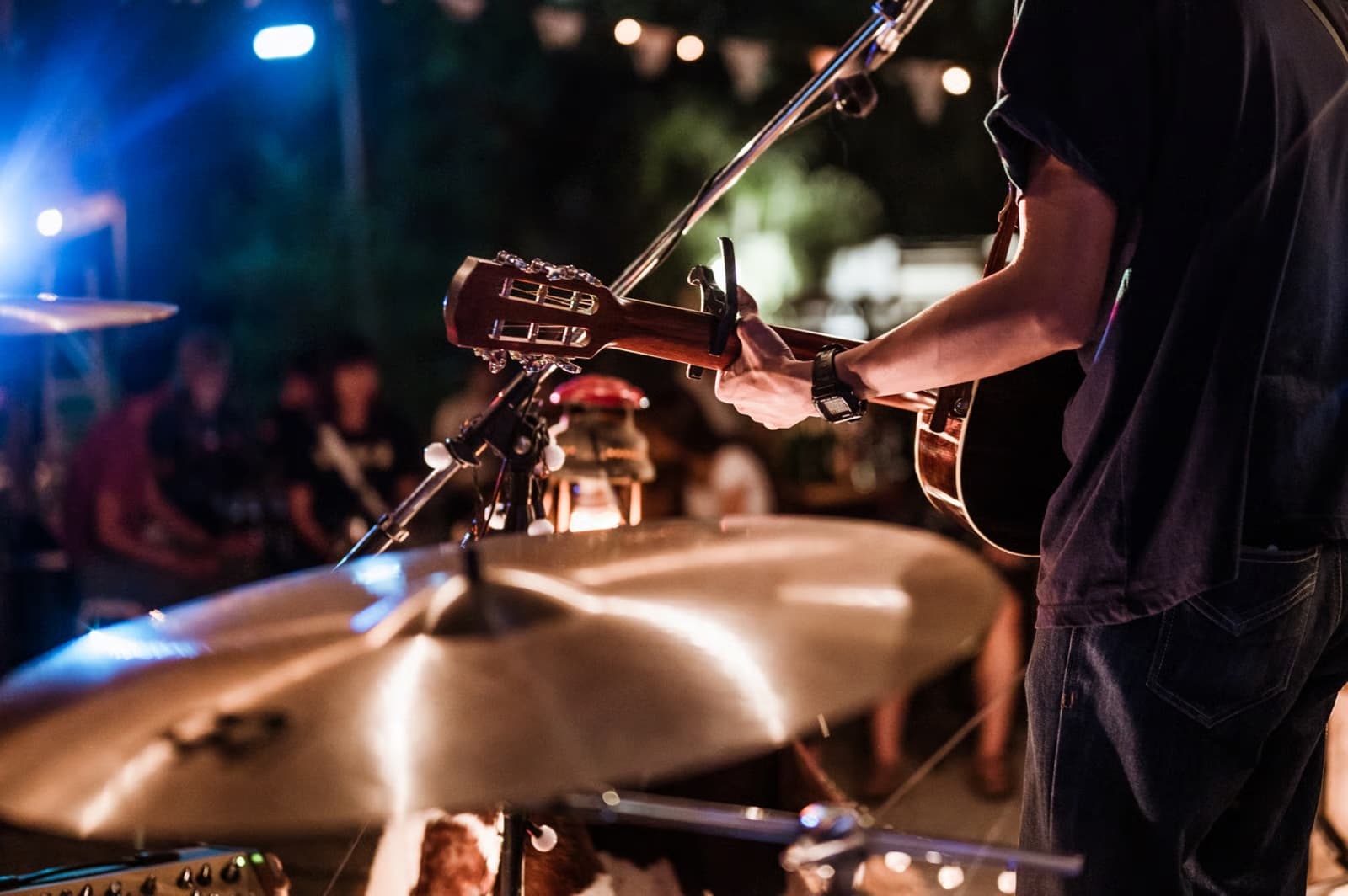 Guitarist playing to a crowd at an outdoor evening event, lit by stage lights and festoon strings