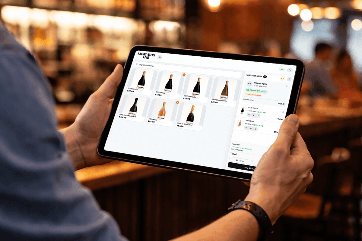 Person holding an iPad displaying the Members One point of sale system in a winery tasting room, with wine bottle listings and order details on screen