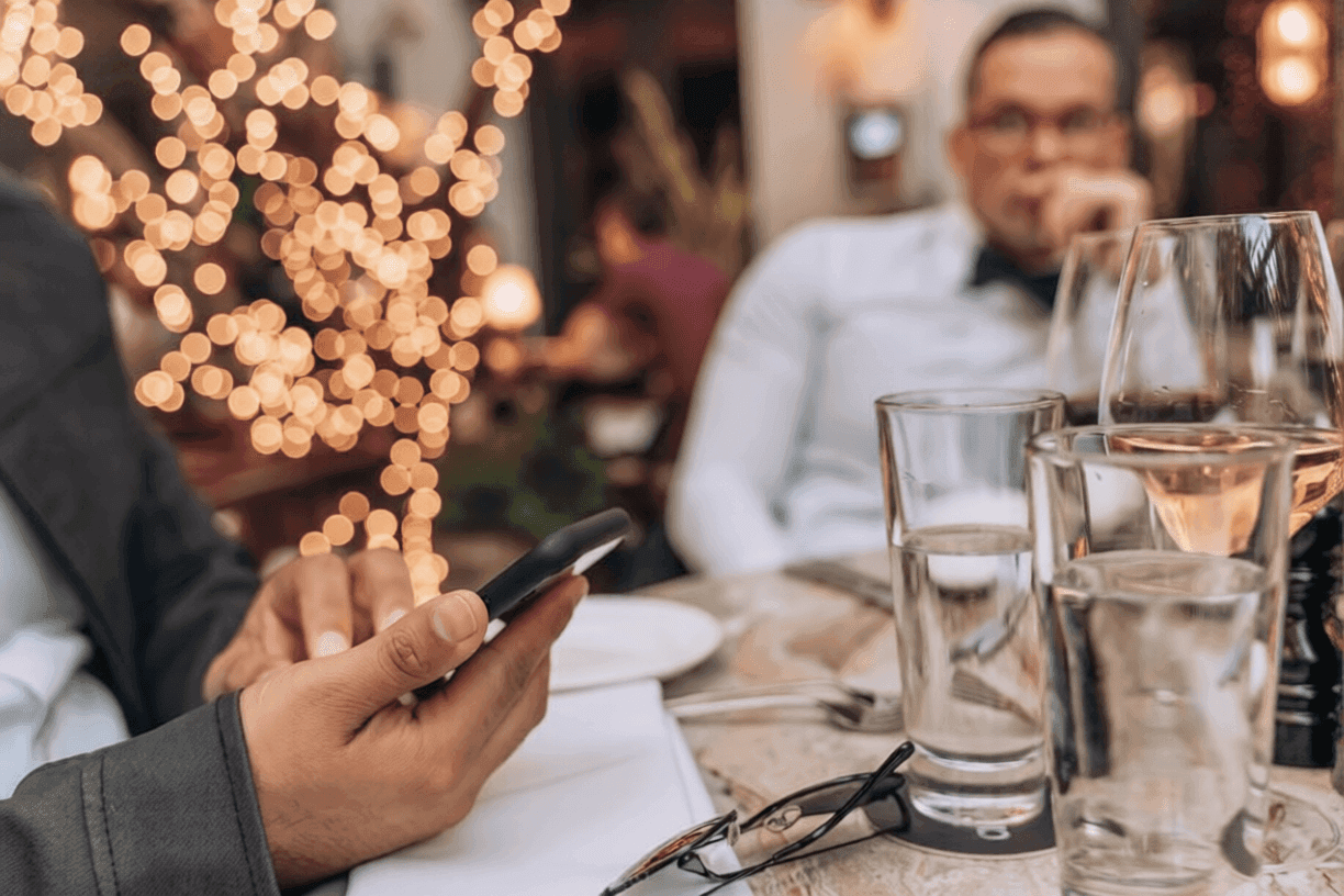 Guest booking a table on their phone at a candlelit restaurant, with wine poured and festoon lights behind them
